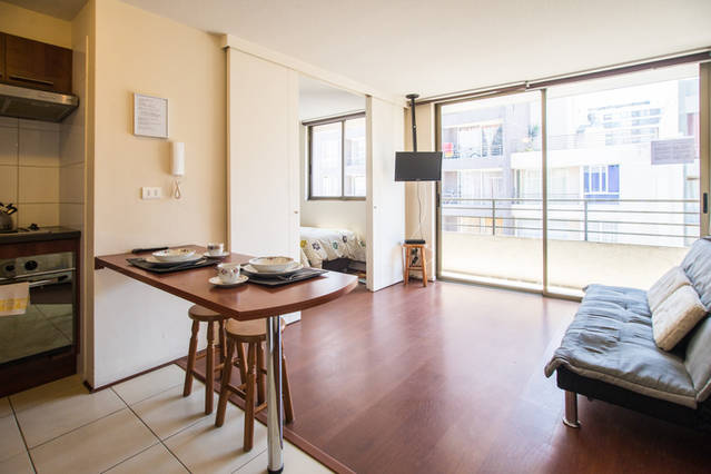 arriendo-departamento-amoblado-palmadesantiago-1402-i departamento con sofa piso de madera televisor ventana mesa con platos y camas de 1 plaza